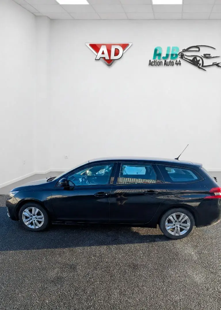 Vue latérale droite d'une Peugeot 308 SW noire stationnée devant un mur blanc avec logos en arrière-plan.