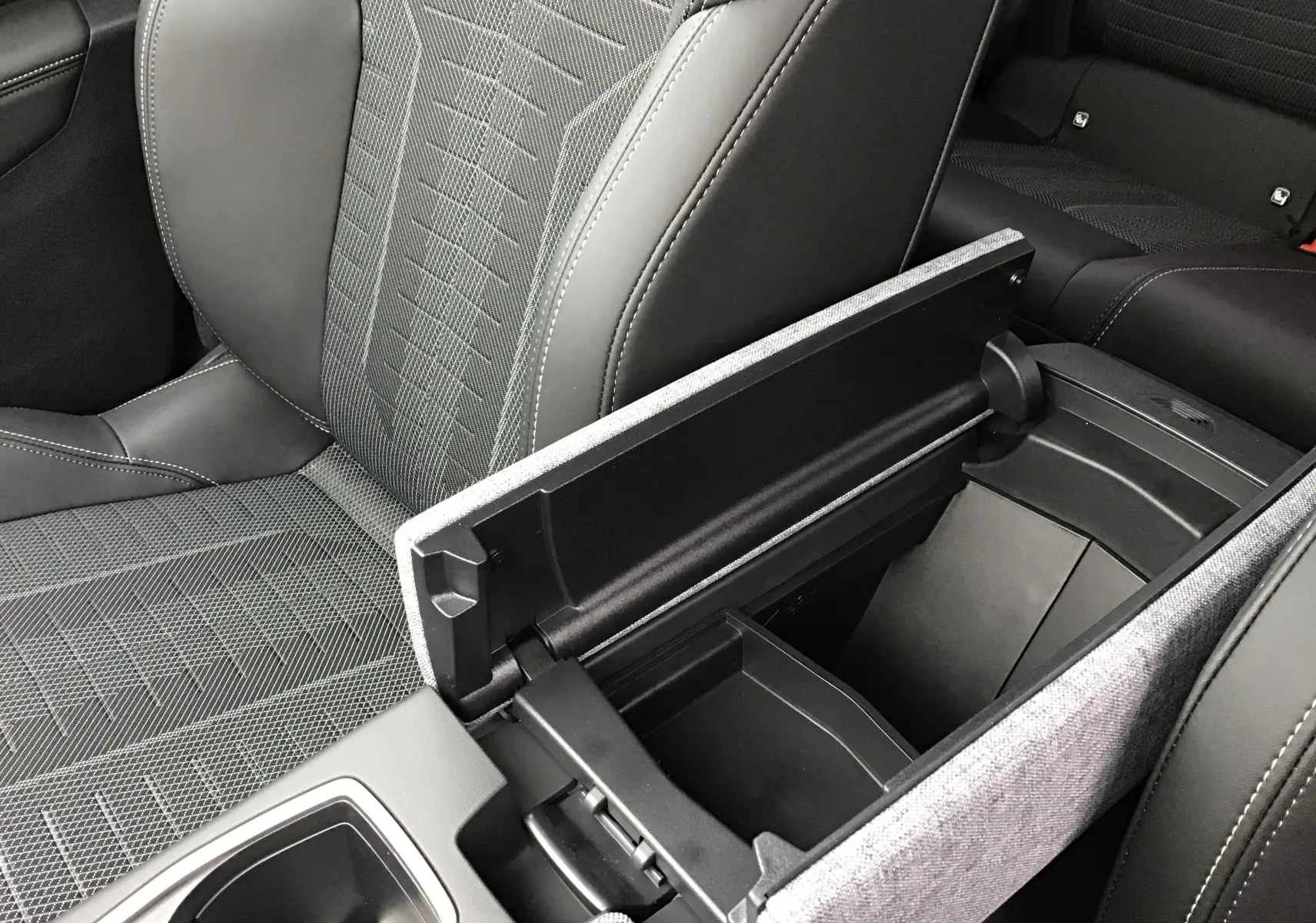 Vue rapprochée de l'accoudoir central ouvert et siège avant en tissu gris et cuir noir dans le Peugeot 3008 hybride 2026.
