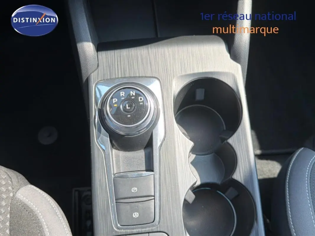 Vue rapprochée du sélecteur rotatif de boîte automatique et porte-gobelets sur la console centrale grise d’une Ford Focus SW bleu foncé.