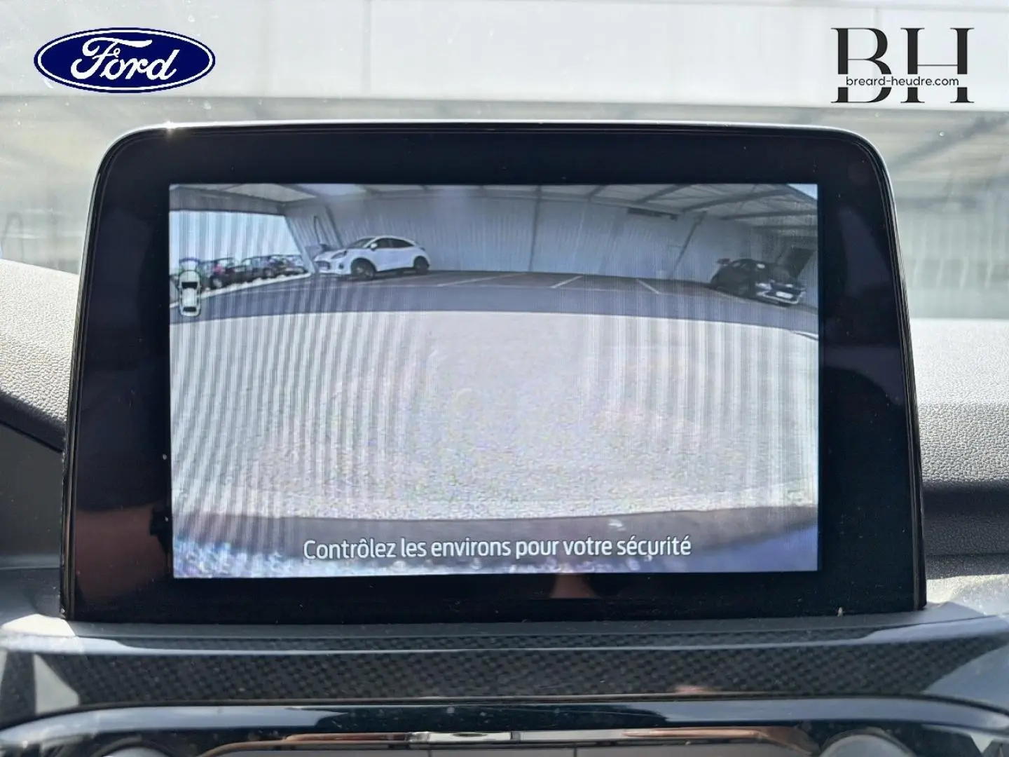 Écran tactile intérieur montrant la caméra de recul du Ford Kuga bleu, avec vue arrière sur parking.