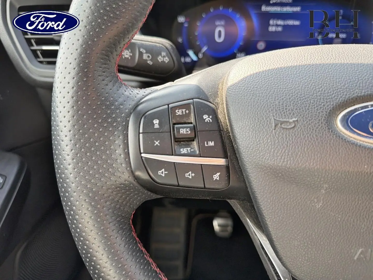 Gros plan sur le volant cuir perforé du Ford Kuga bleu, avec commandes multifonctions et tableau de bord numérique en arrière-plan.