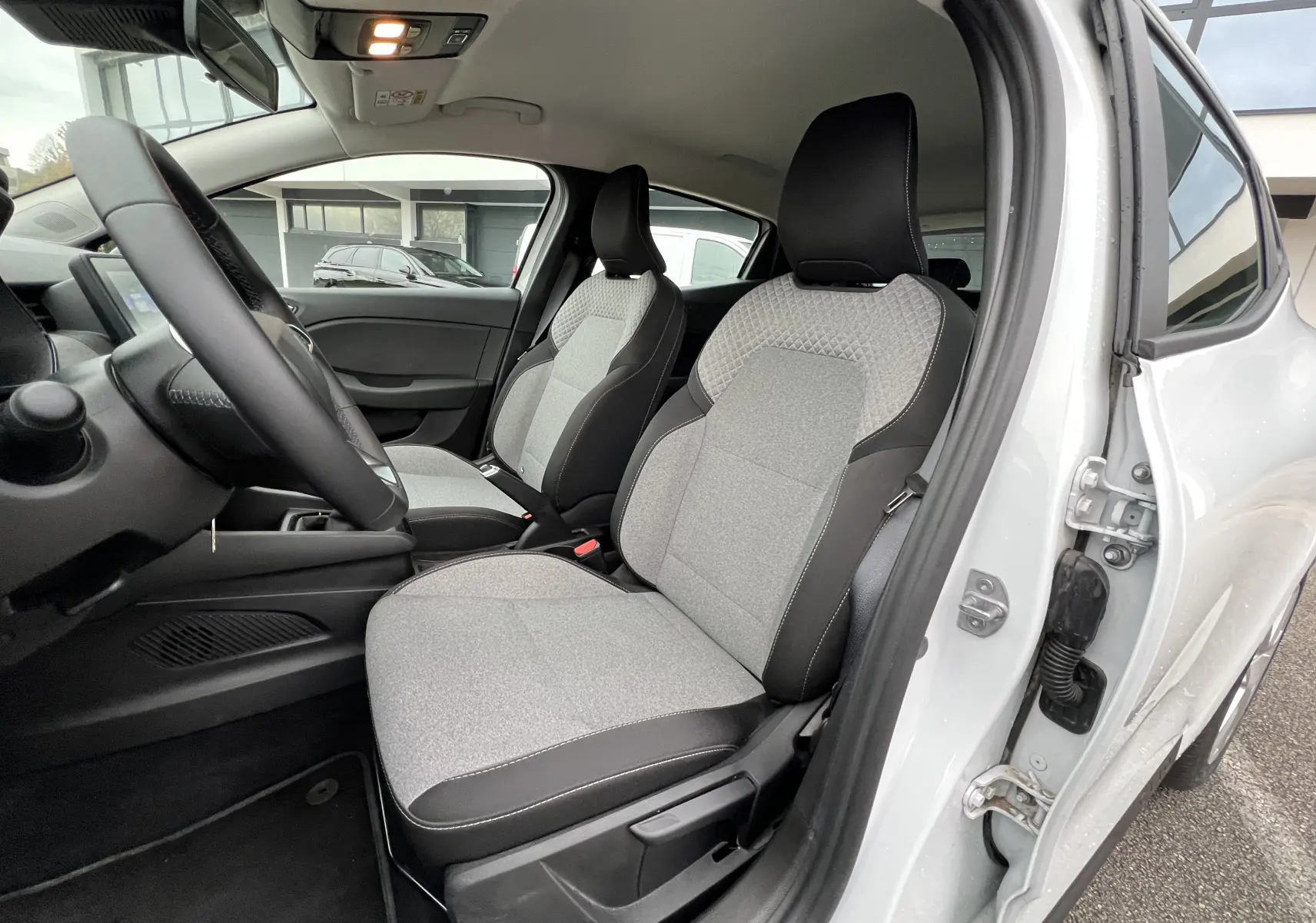 Vue intérieure côté conducteur de la Renault Clio V blanche 2025 avec sièges tissu gris et volant cuir.