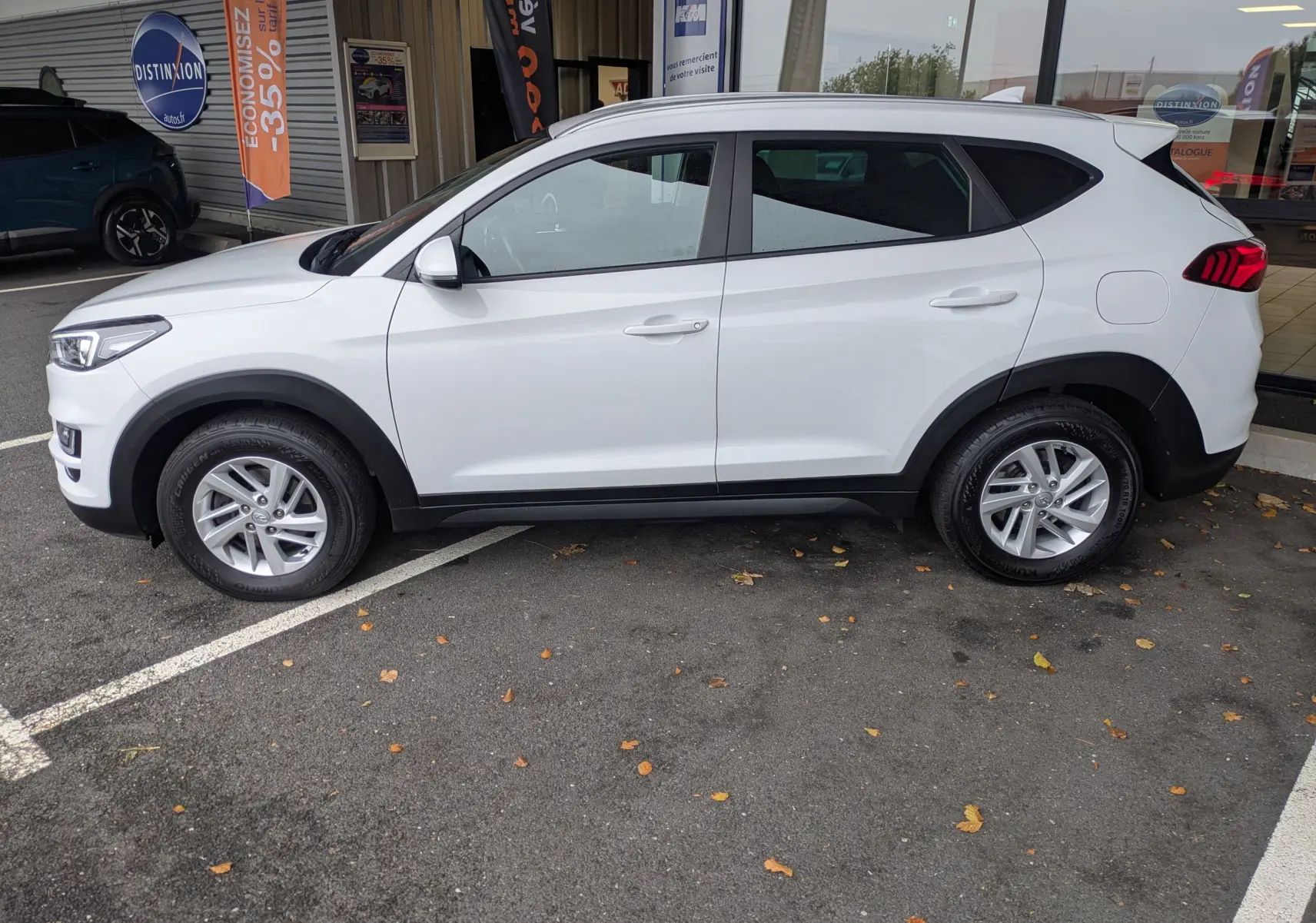 Vue de profil côté gauche d'un Hyundai Tucson blanc 2020 stationné devant un showroom avec jantes alliage et feux arrière LED.