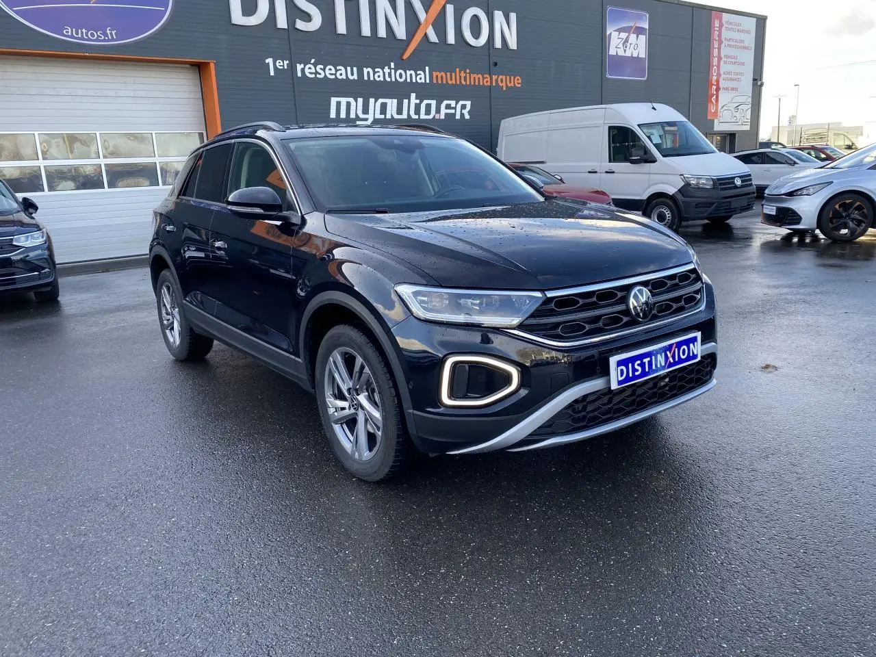 Volkswagen T-Roc noir nacré en 3/4 avant droit, avec feux LED et calandre distinctive sur parking humide.