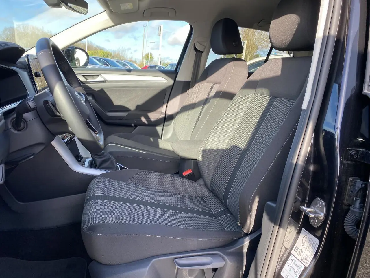 Vue intérieure côté conducteur du Volkswagen T-Roc noir 2025, montrant les sièges avant en tissu gris et le tableau de bord.