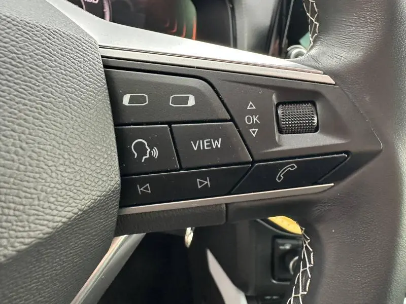 Gros plan sur les commandes à gauche du volant du SEAT Arona 2025, boutons noirs avec marquages blancs sur cuir noir.