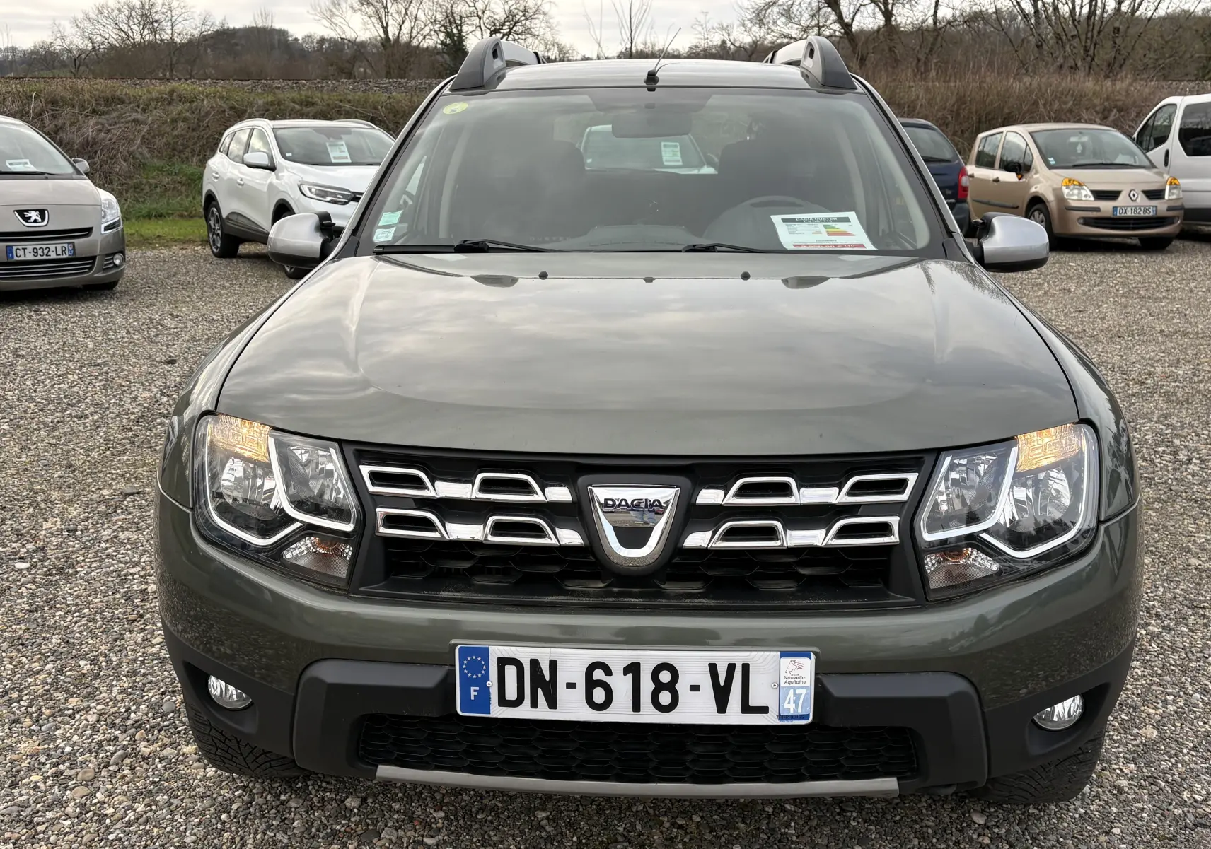 Vue de face d’un Dacia Duster 1.5 DCI 110 vert foncé avec calandre chromée et phares allumés sur un parking.