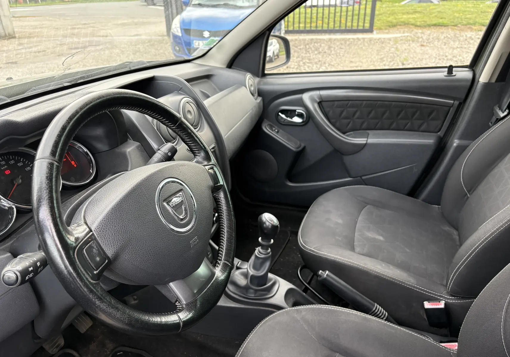 Vue intérieure côté conducteur du Dacia Duster 2014, montrant le volant, la boîte manuelle et les sièges noirs.