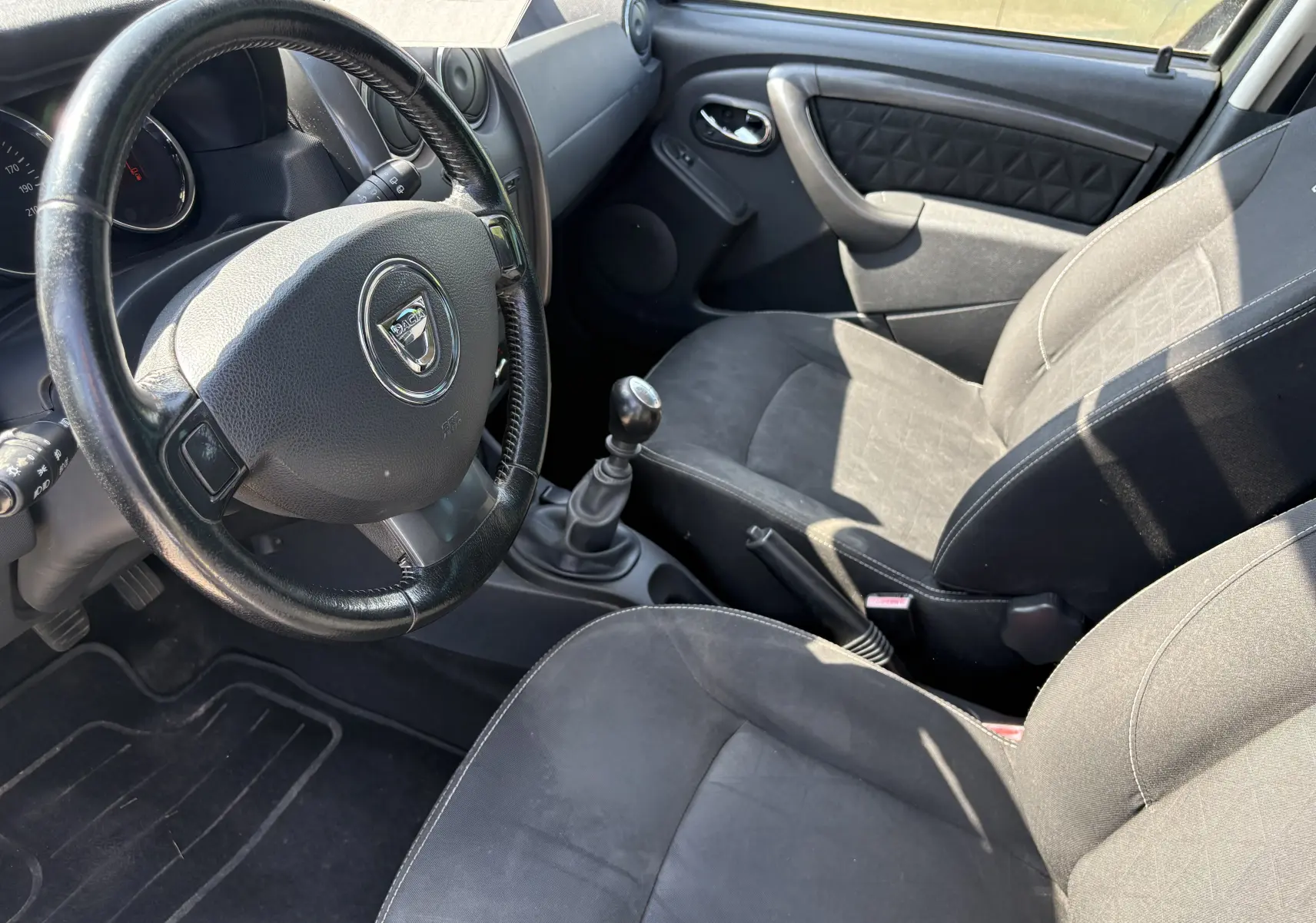 Vue intérieure côté conducteur du Dacia Duster 1.5 DCI 110 de 2014, volant cuir, sièges tissu gris foncé et levier manuel.
