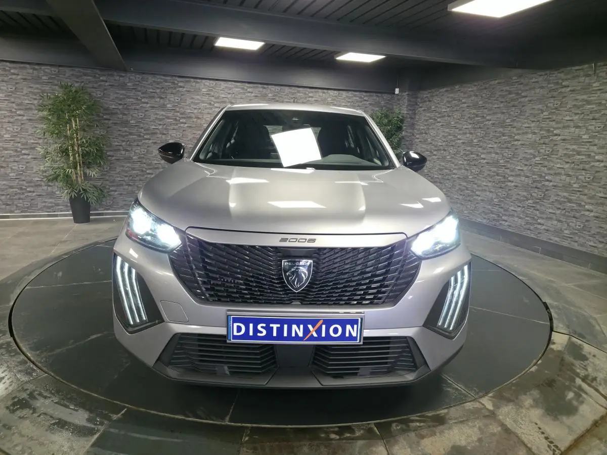 Vue de face du Peugeot 2008 gris métallisé 2025 avec calandre noire et feux LED allumés dans un showroom.