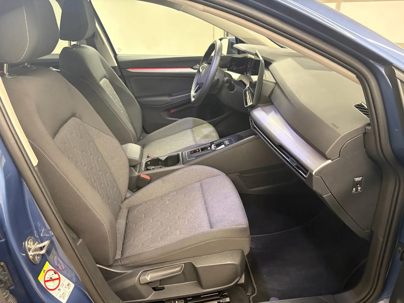 Vue intérieure côté conducteur de la Volkswagen Golf 1.5 eTSI 2025 en Bleu Anémone, sièges tissu gris et tableau de bord moderne.