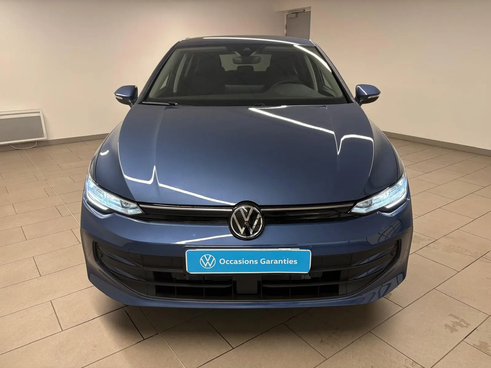 Vue frontale d'une Volkswagen Golf 1.5 eTSI 116 DSG7 VW Edition en bleu Anémone avec phares allumés dans un showroom.