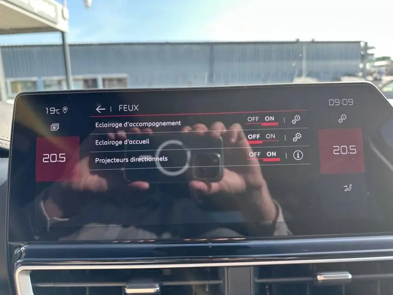 Écran tactile intérieur du Citroën C5 Aircross 2025 affichant les réglages des feux, avec reflet de la main et appareil photo.