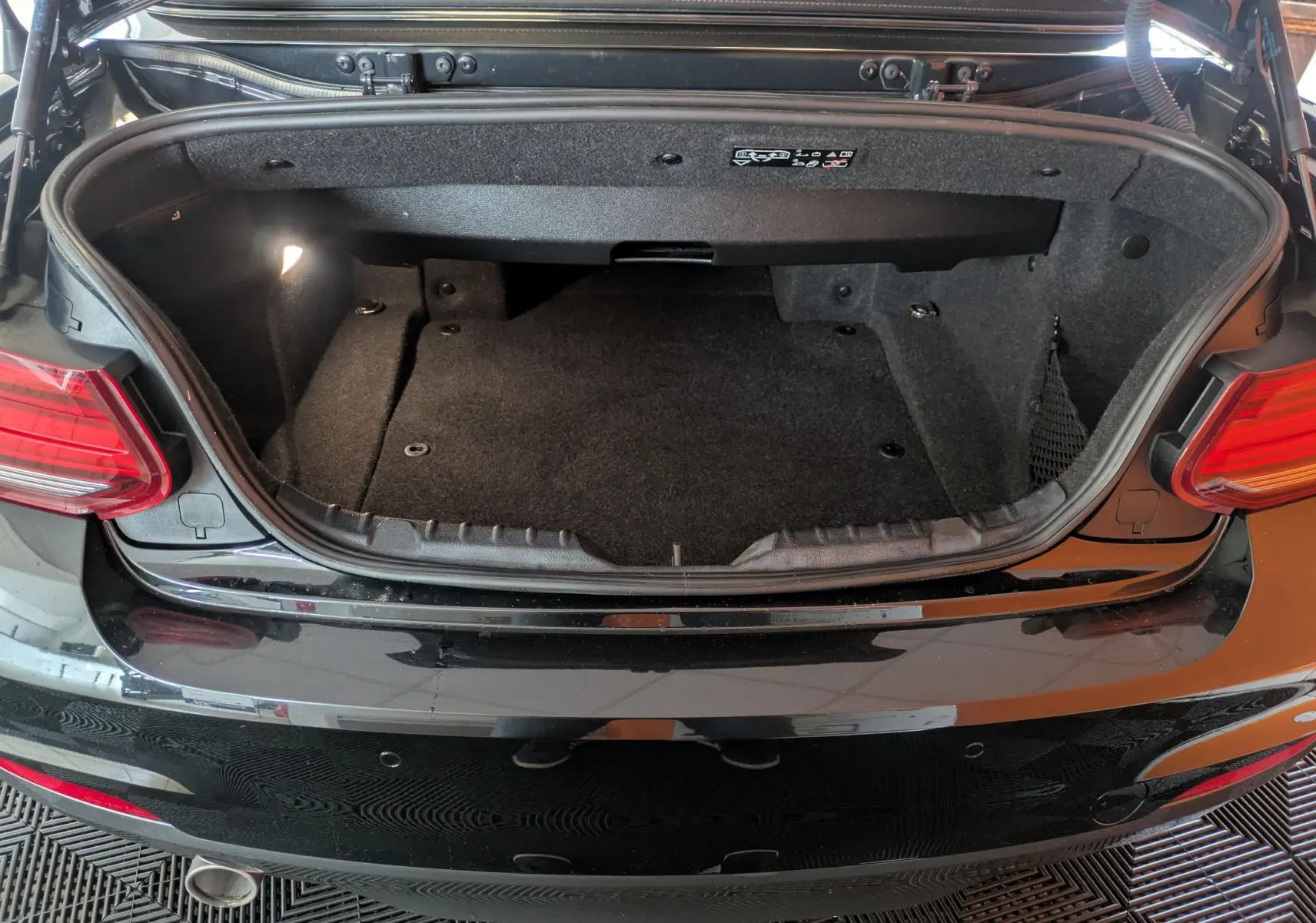Coffre ouvert vu de l'arrière d'une BMW Série 2 Cabriolet 218d noire, montrant l'espace de rangement et l'éclairage intérieur.