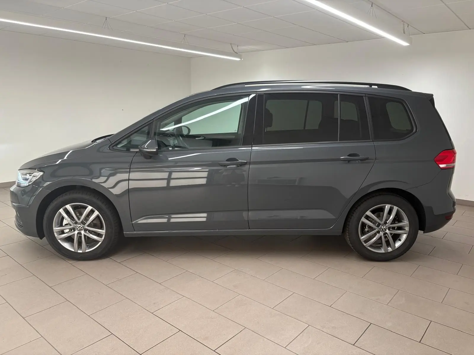 Profil côté gauche d'un Volkswagen Touran gris dauphin 2025 avec jantes alliage et lignes épurées dans un intérieur lumineux.
