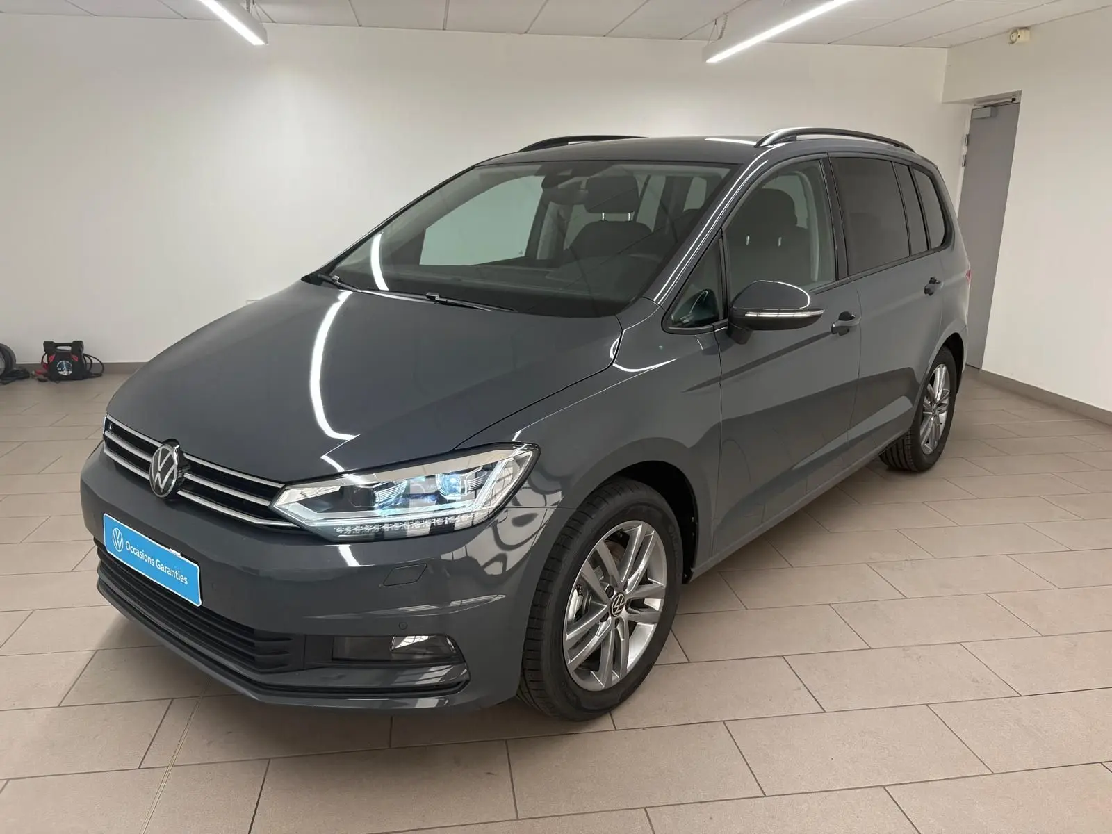 Volkswagen Touran gris dauphin en 3/4 avant droit dans un showroom, avec jantes alliage et feux LED allumés.
