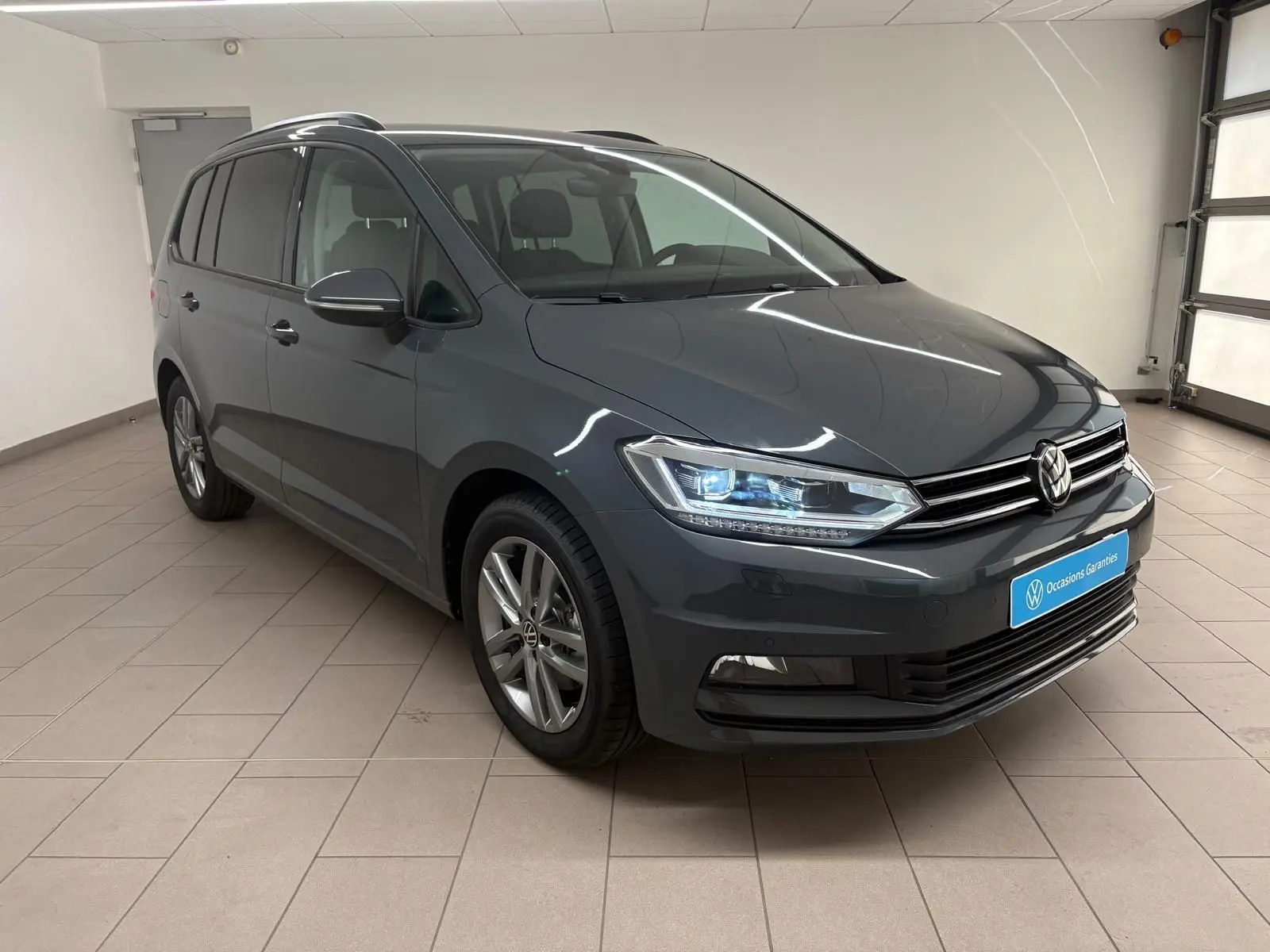 Volkswagen Touran gris dauphin en 3/4 avant droit, avec phares LED allumés et jantes alliage 17 pouces.
