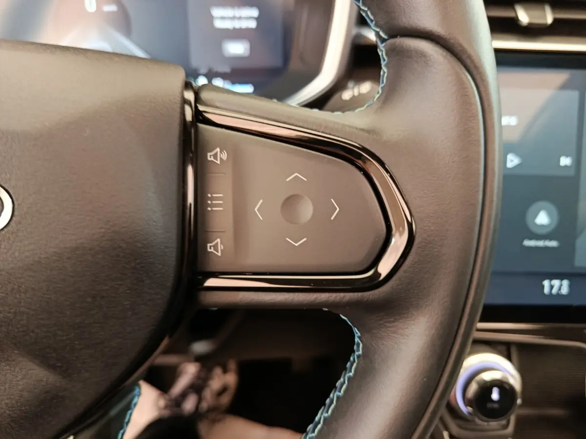 Gros plan sur les commandes tactiles du volant en cuir noir du Link & Co 01 hybride rechargeable, avec écran tactile en arrière-plan.