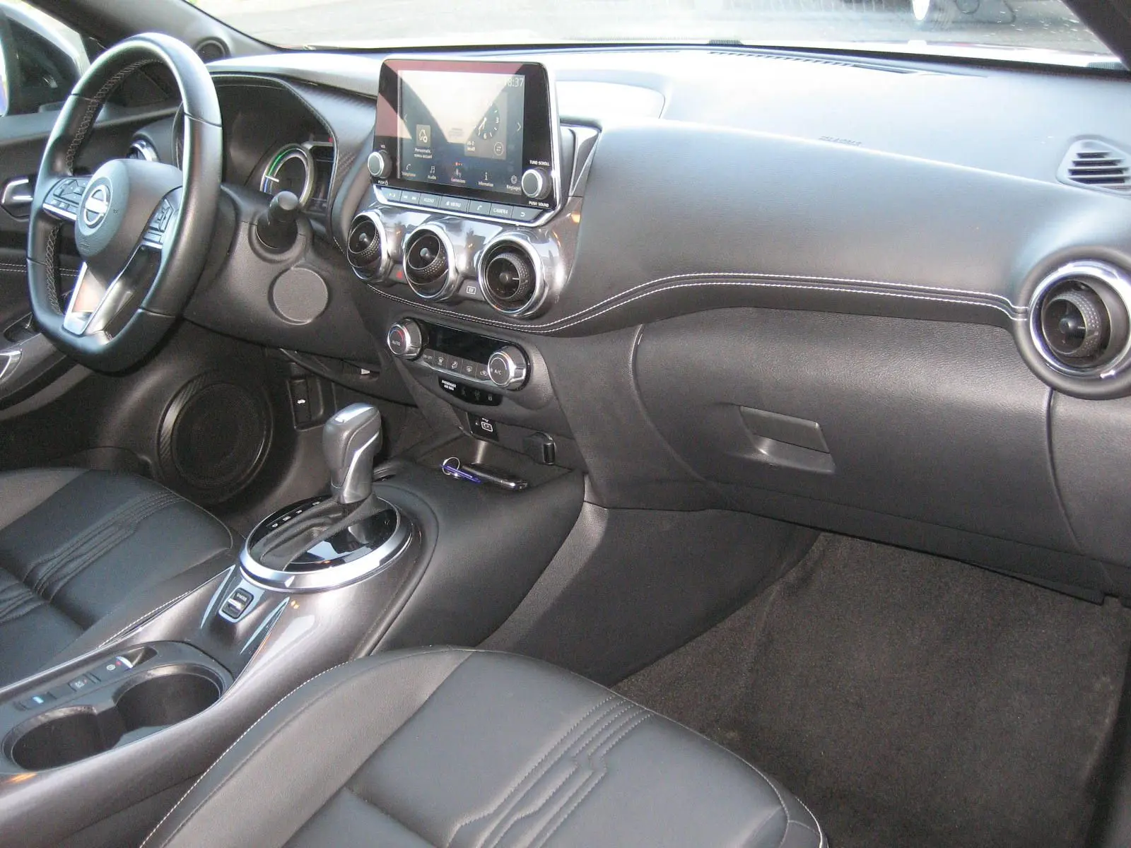 Vue intérieure côté conducteur du tableau de bord et console centrale du Nissan Juke 1.6 Hybrid 143 Tekna avec sellerie cuir noire.