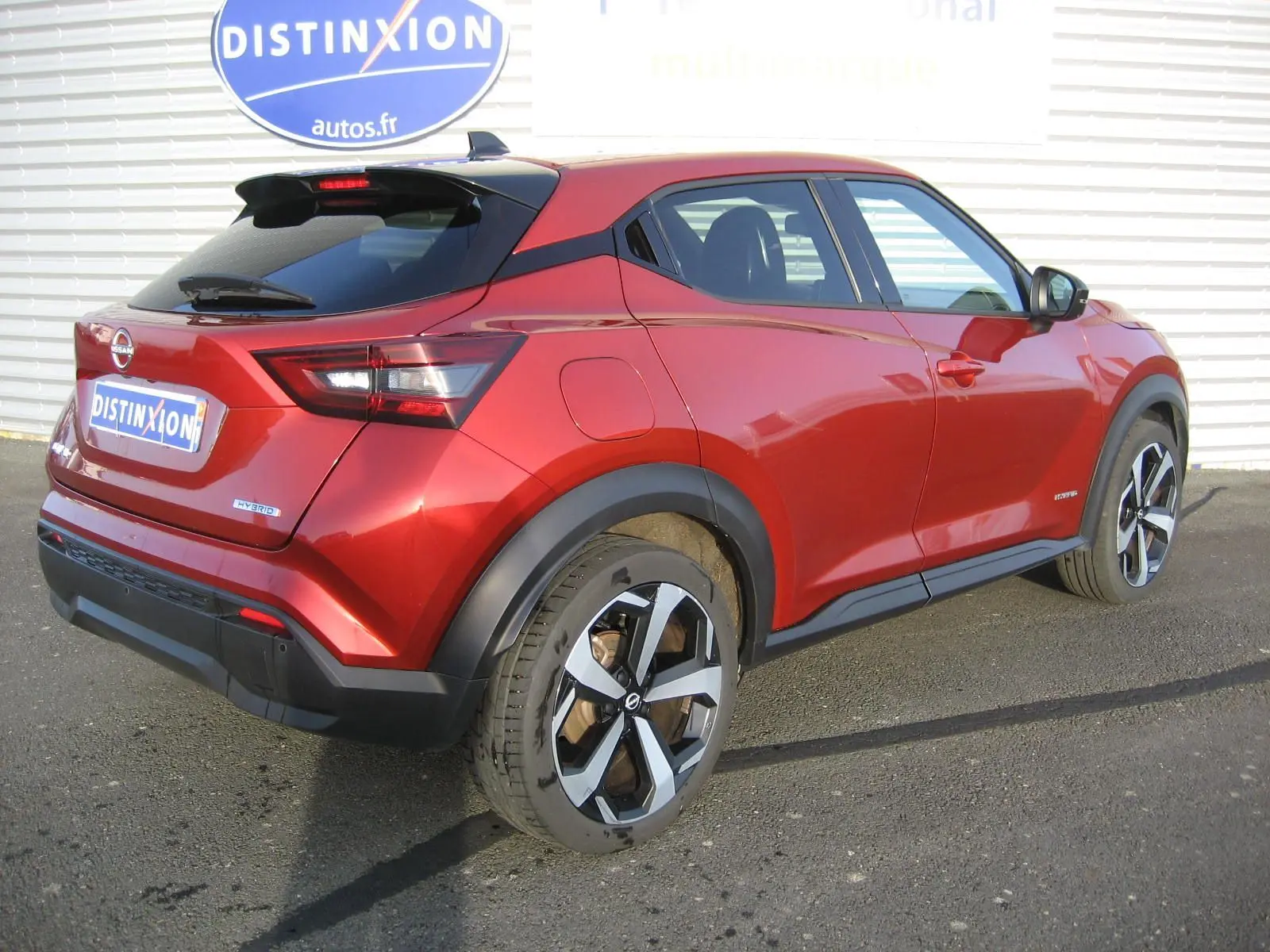 Vue 3/4 arrière droite d'un Nissan Juke 1.6 Hybrid rouge 2024 avec jantes bi-ton et toit noir.