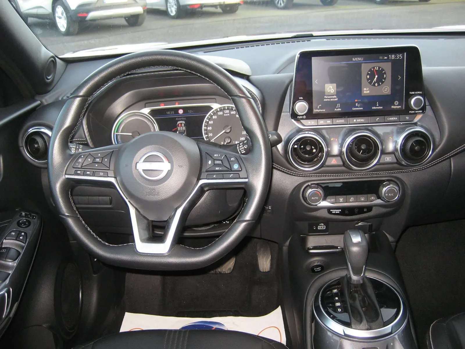 Vue intérieure centrée sur le volant et la console centrale du Nissan Juke 1.6 Hybrid rouge avec écran tactile et boîte auto.