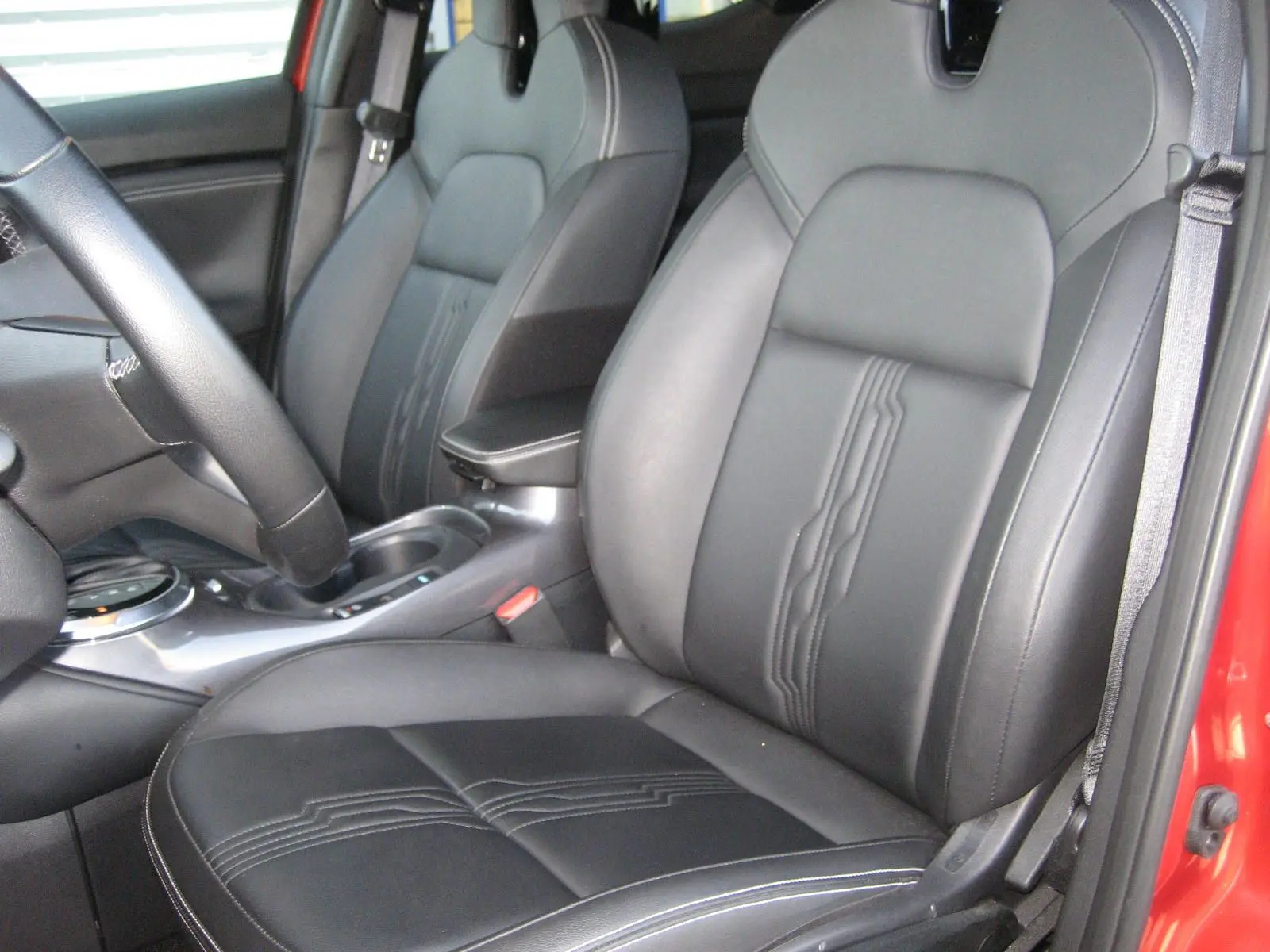 Vue intérieure côté conducteur du Nissan Juke rouge 2024, mettant en valeur les sièges en cuir noir avec surpiqûres décoratives.