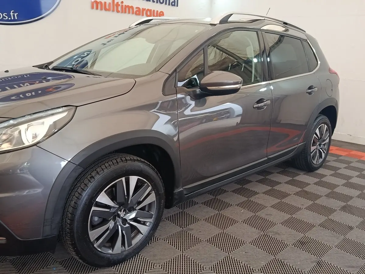 Vue 3/4 avant droit d’un Peugeot 2008 gris foncé avec jantes alliage et barres de toit chromées en intérieur showroom.