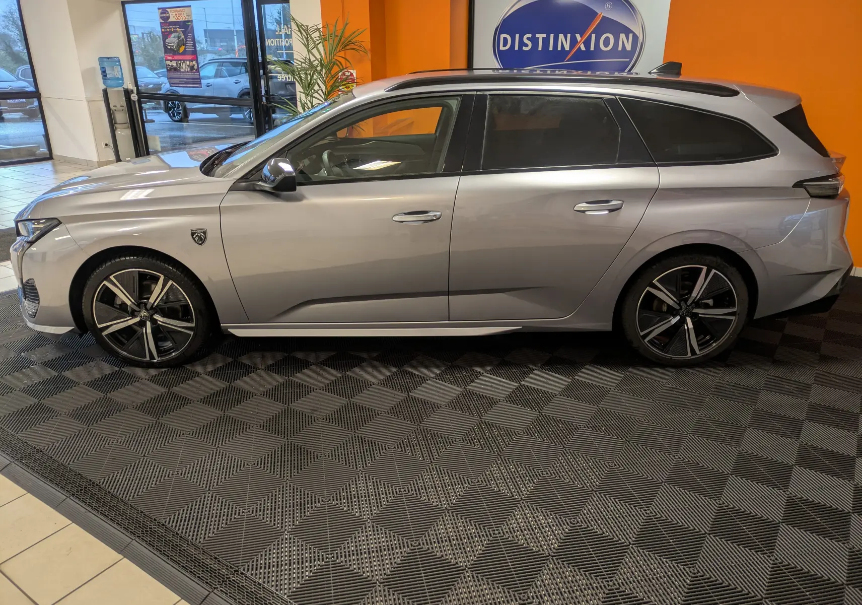 Vue latérale droite d’un Peugeot 308 SW Hybrid gris 2025 en showroom, avec jantes alliage 18 pouces et toit ouvrant.