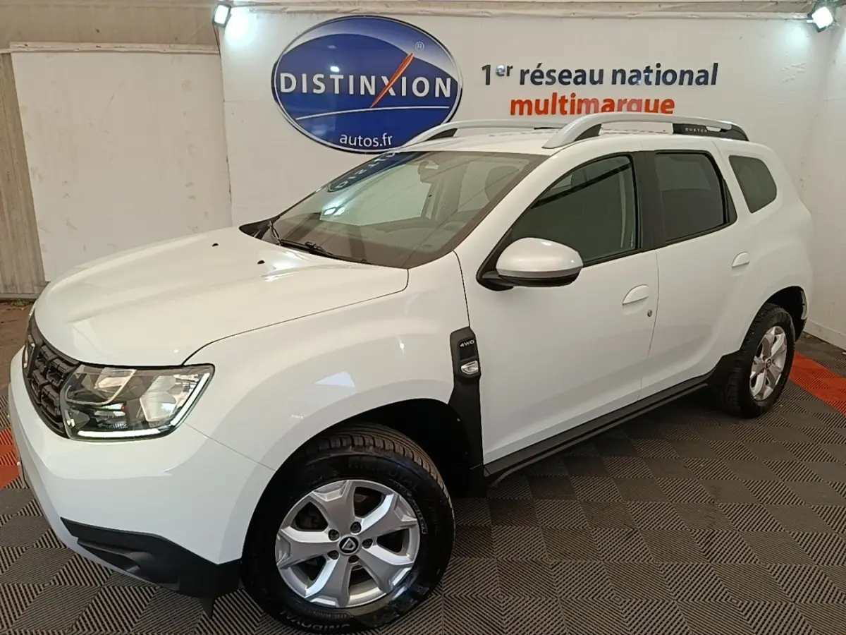SUV Dacia Duster blanc en 3/4 avant droit, avec barres de toit noires et jantes alliage visibles.