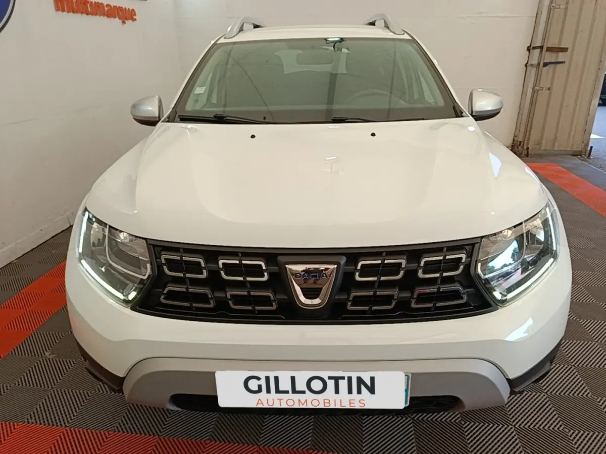 Vue avant d'un Dacia Duster blanc avec calandre noire et phares LED allumés en intérieur showroom.