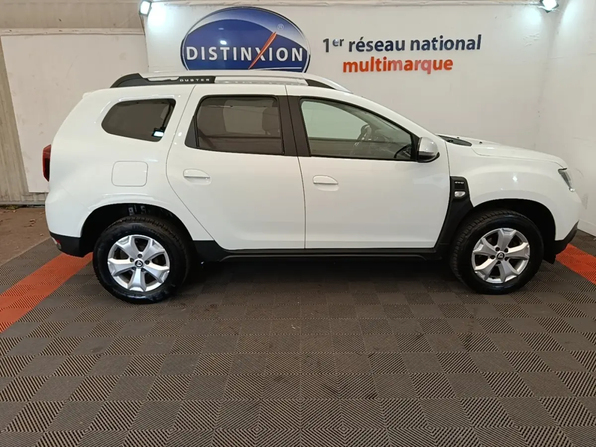 Vue de profil côté droit du Dacia Duster blanc Confort Blue dCi 115 4x4 avec rails de toit noirs en intérieur showroom.