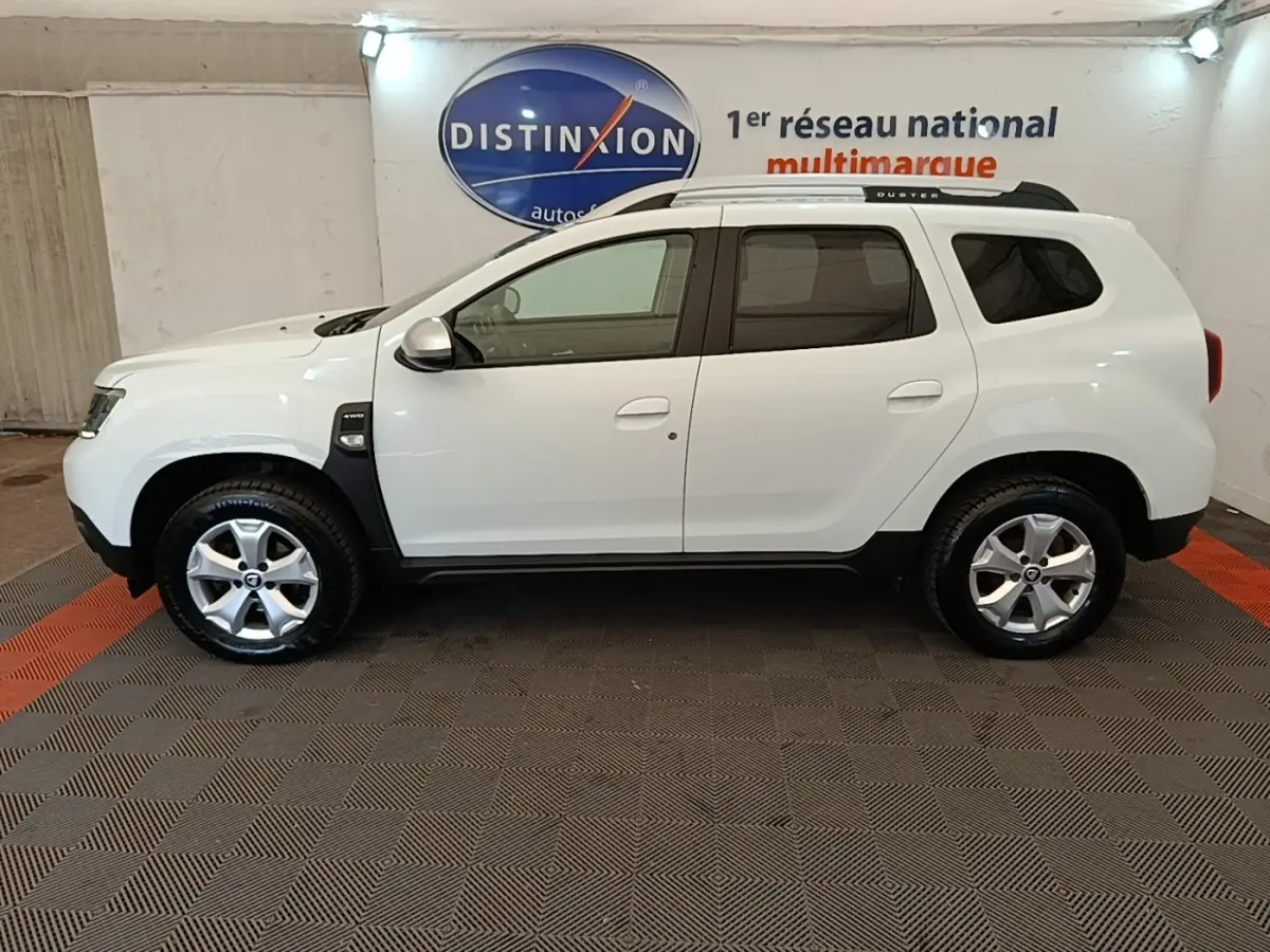 Vue de profil droit du Dacia Duster blanc 2021 avec barres de toit noires et jantes alliage en intérieur showroom.