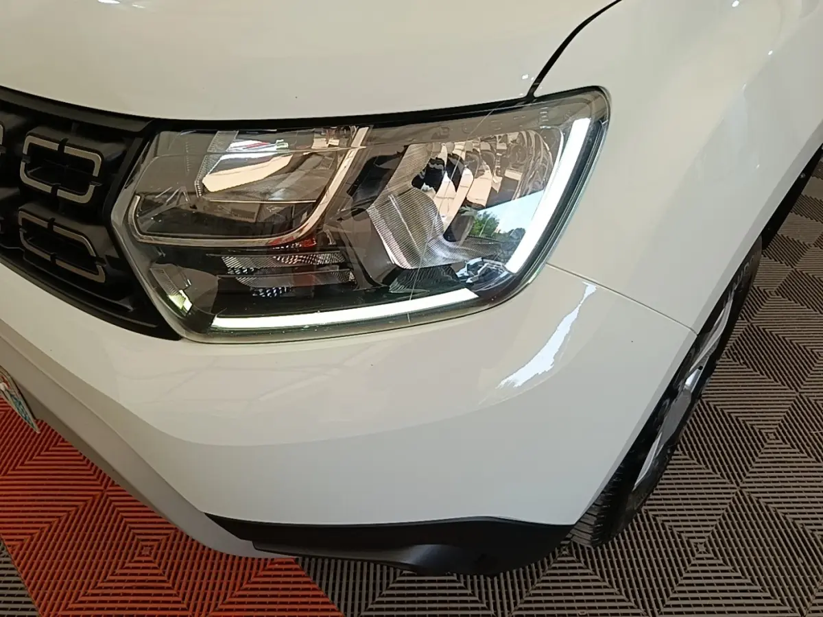 Gros plan sur le phare avant droit du Dacia Duster blanc, soulignant la calandre noire et le pare-chocs gris.