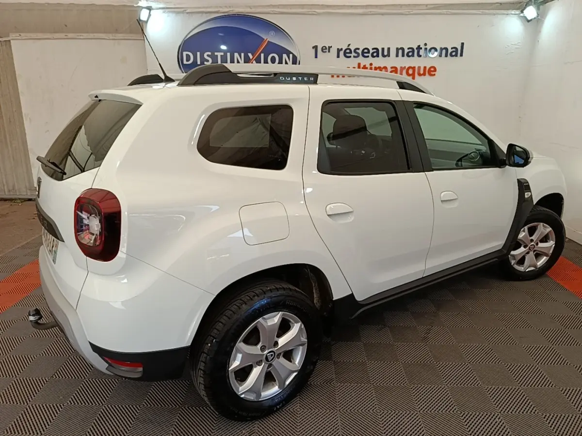 Vue 3/4 arrière droite d'un Dacia Duster blanc 2021 avec barres de toit noires et jantes alliage.