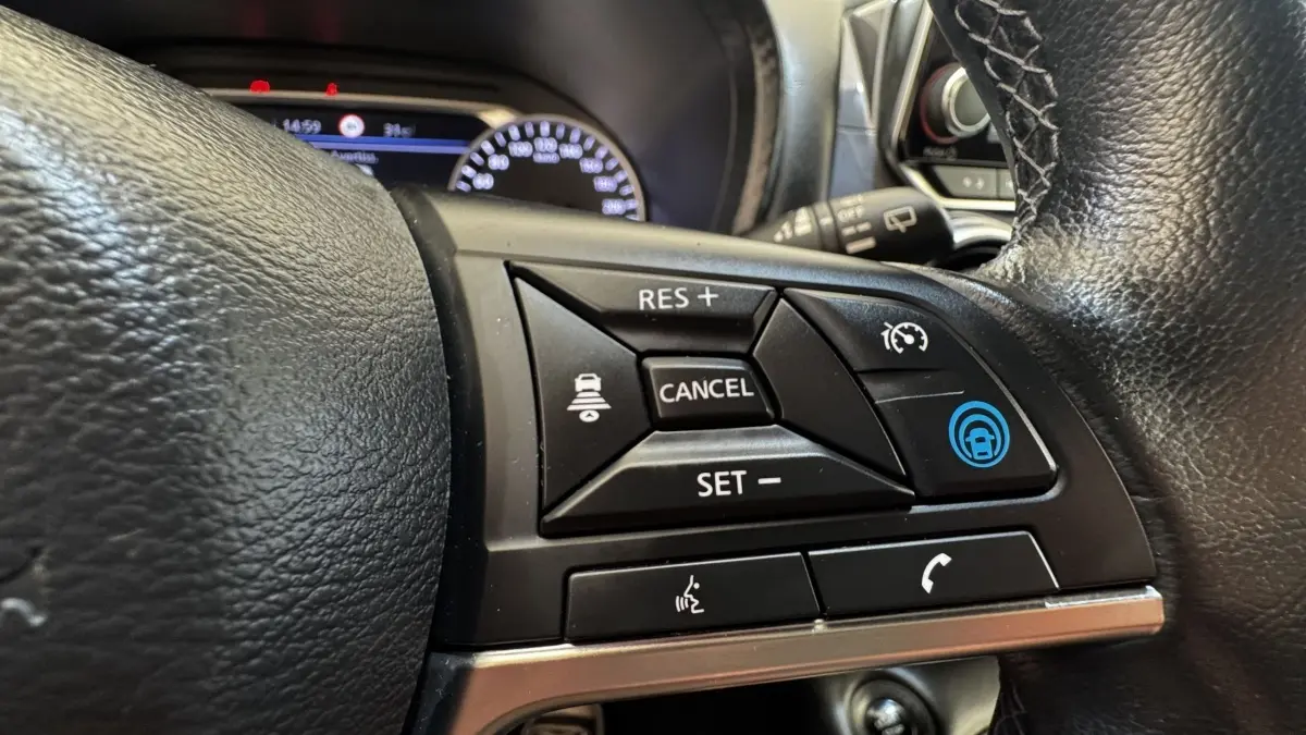 Gros plan sur les commandes du volant noir du Nissan Juke Hybrid 143 Business+ 2023, affichage du combiné en arrière-plan.