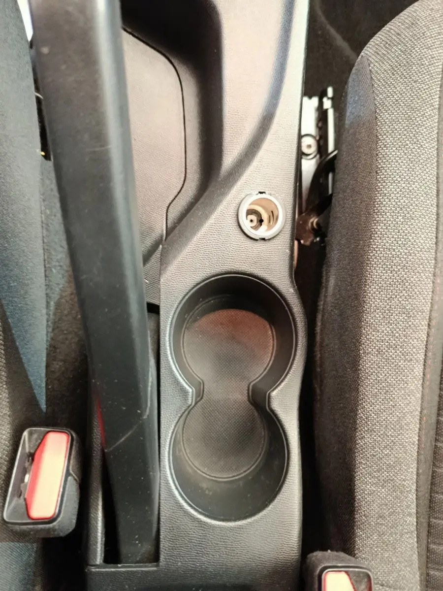 Gros plan sur la console centrale noire avec porte-gobelets et prise 12V entre les sièges avant gris foncé.