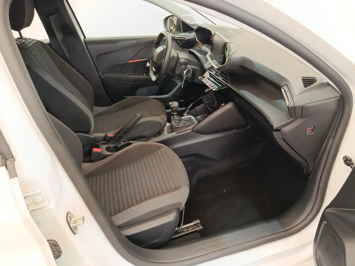 Intérieur avant droit de la Peugeot 208 blanche, montrant sièges tissu noirs et tableau de bord avec écran tactile.