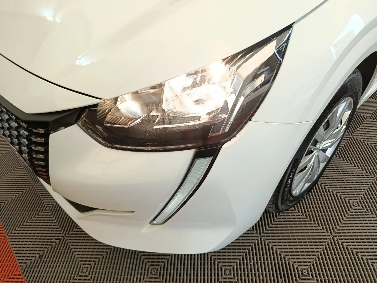 Gros plan sur le phare avant droit allumé d'une Peugeot 208 blanche, avec la calandre et la roue visibles.