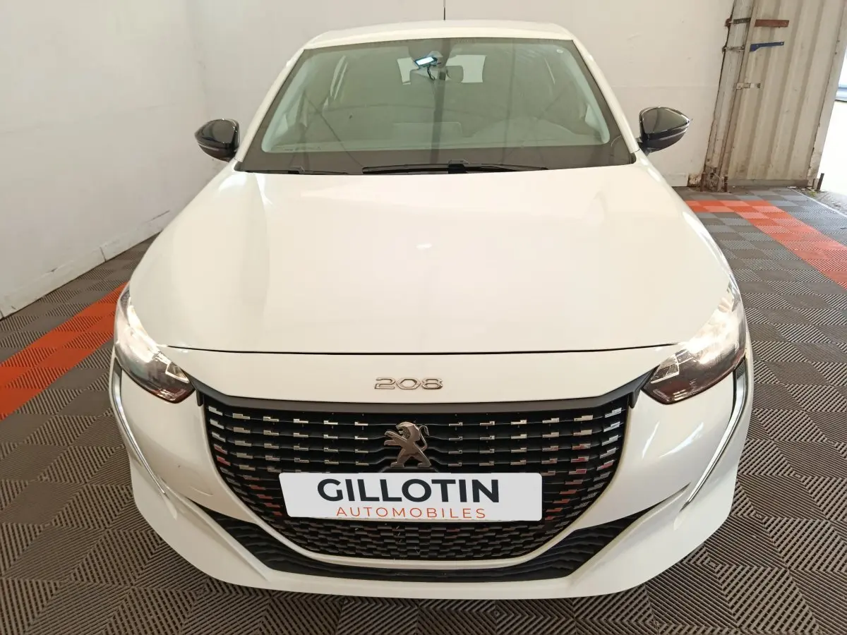 Vue avant d'une Peugeot 208 blanche 2023 avec calandre noire et phares allumés dans un garage.