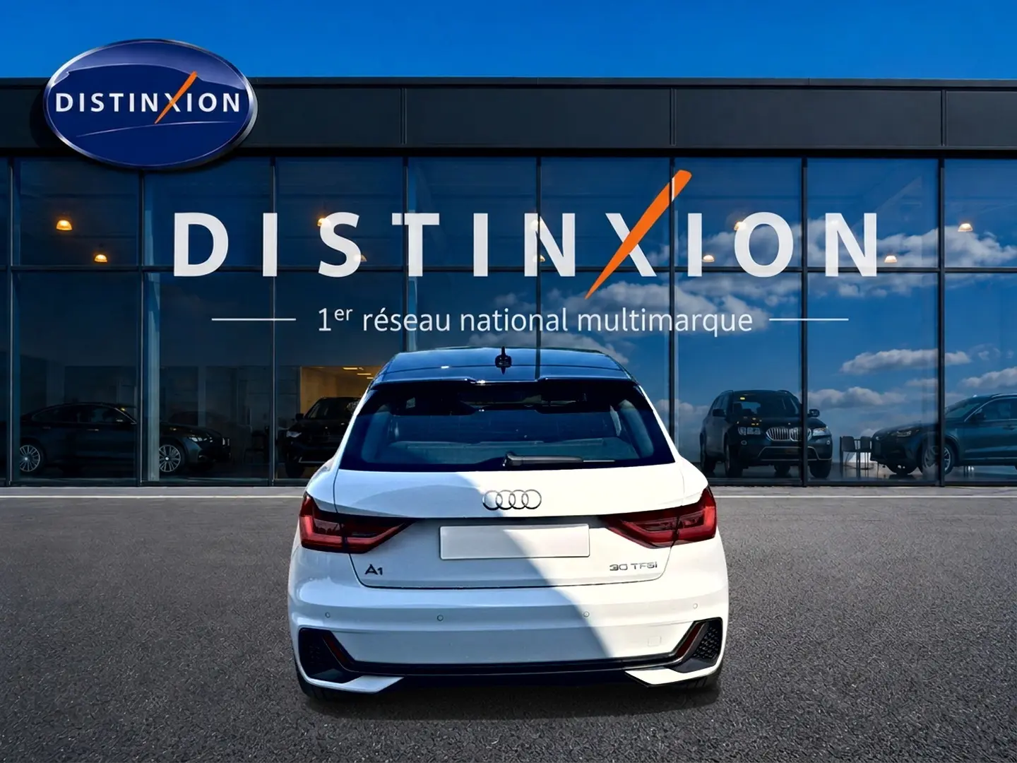 Vue arrière d'une Audi A1 Sportback blanche avec toit noir, feux LED et becquet arrière, stationnée devant un showroom Distinxion.