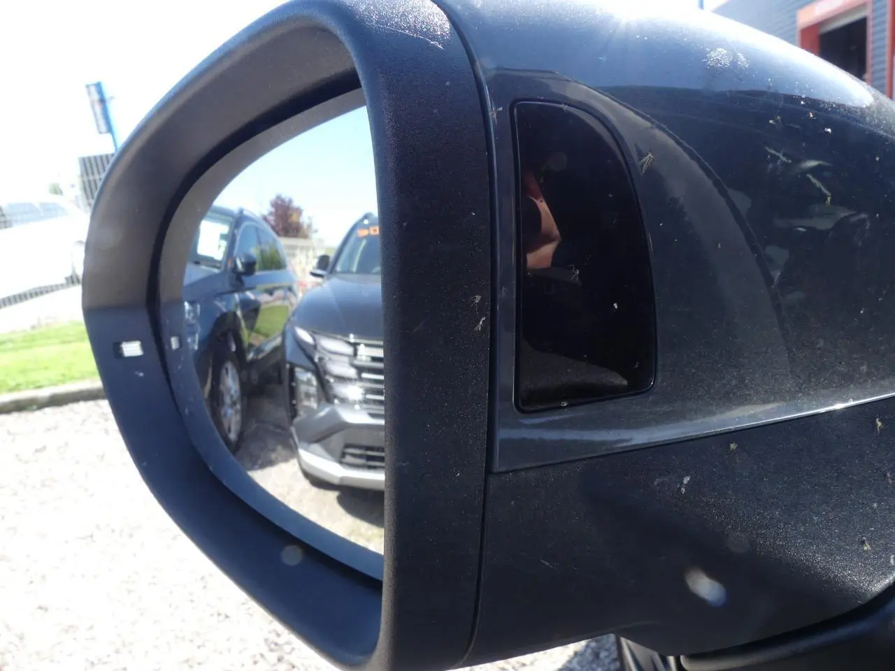 Rétroviseur extérieur droit noir avec reflet d'un SUV gris foncé garé à côté sur un parking.