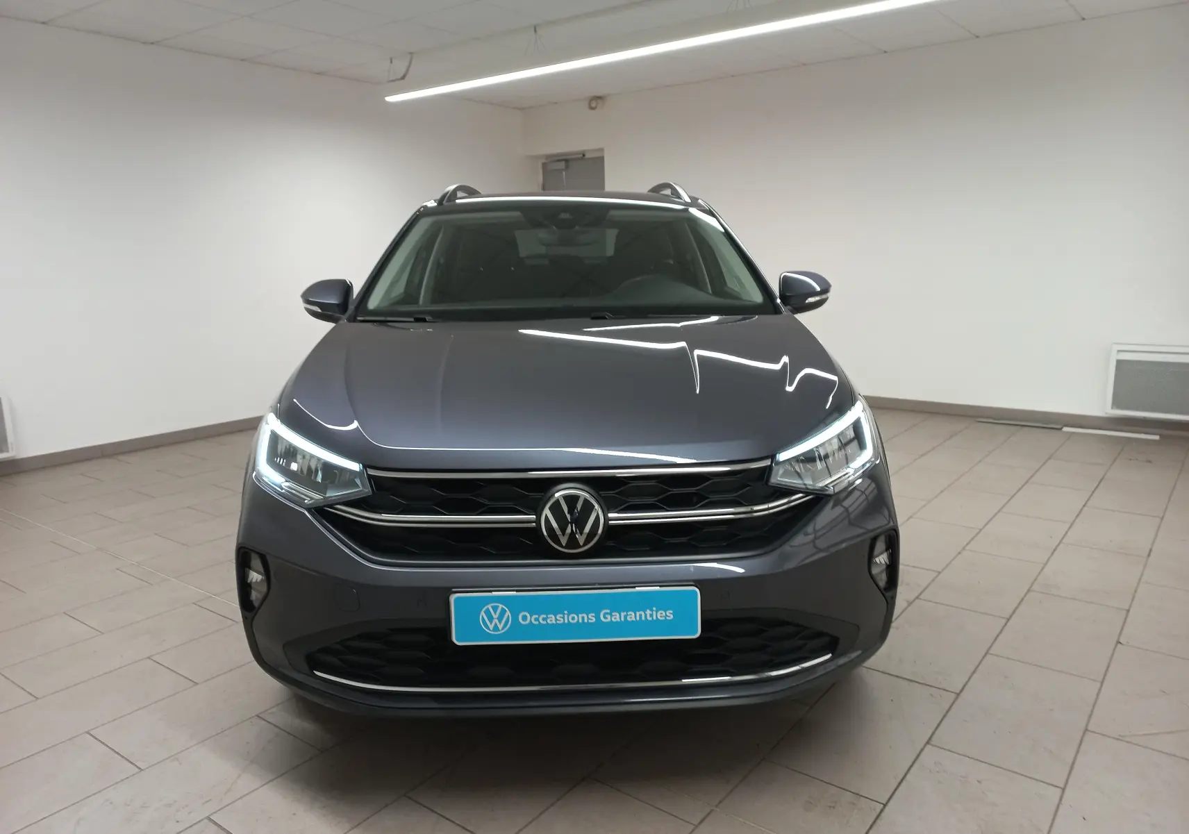 Vue avant du Volkswagen Taigo 1.0 TSI 116 DSG7 Gris Cendré avec projecteurs LED et calandre noire en intérieur showroom.