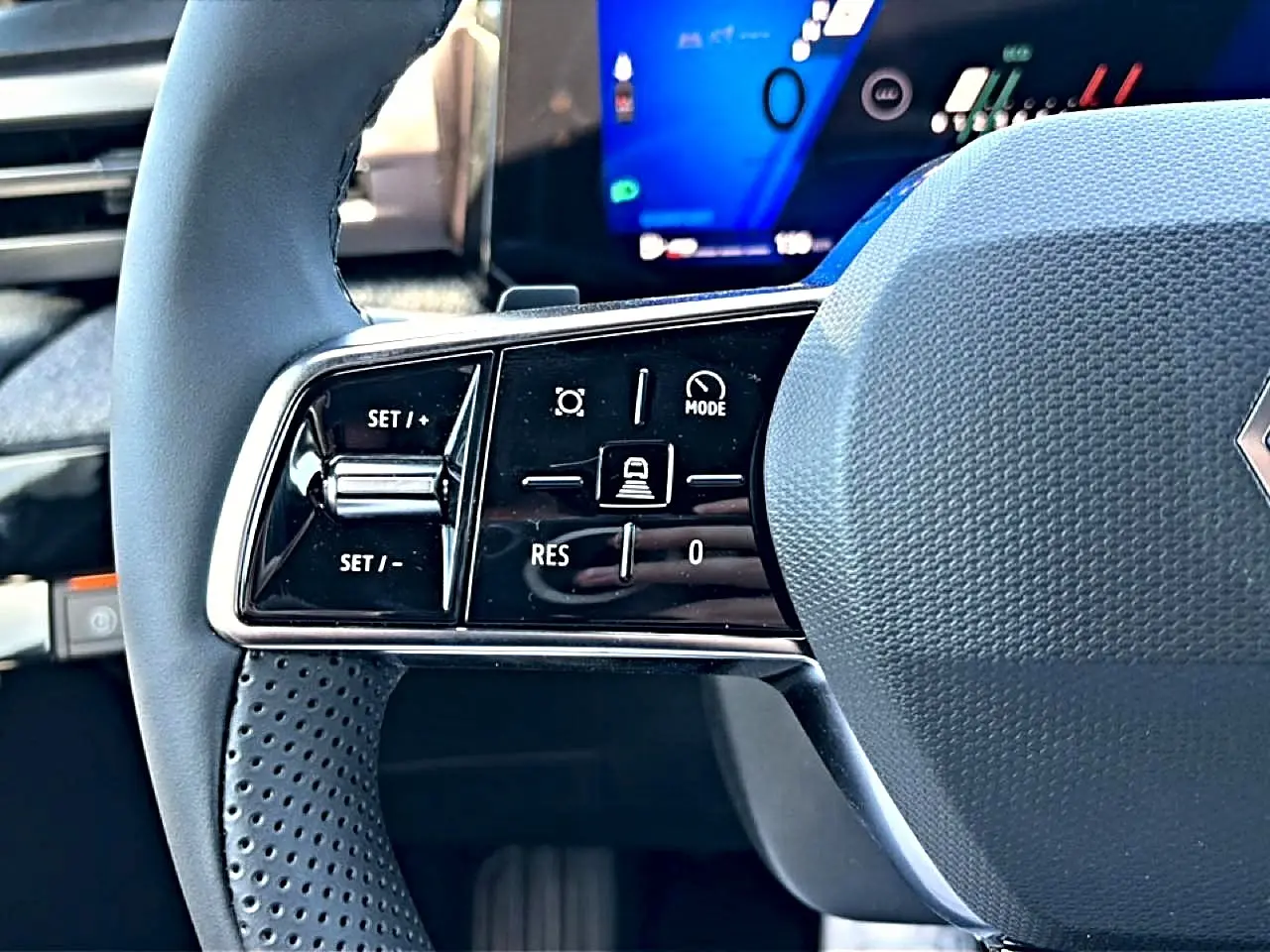 Gros plan sur la partie gauche du volant gris de la Renault Clio VI avec commandes de régulateur de vitesse et écran digital flou en arrière-plan.