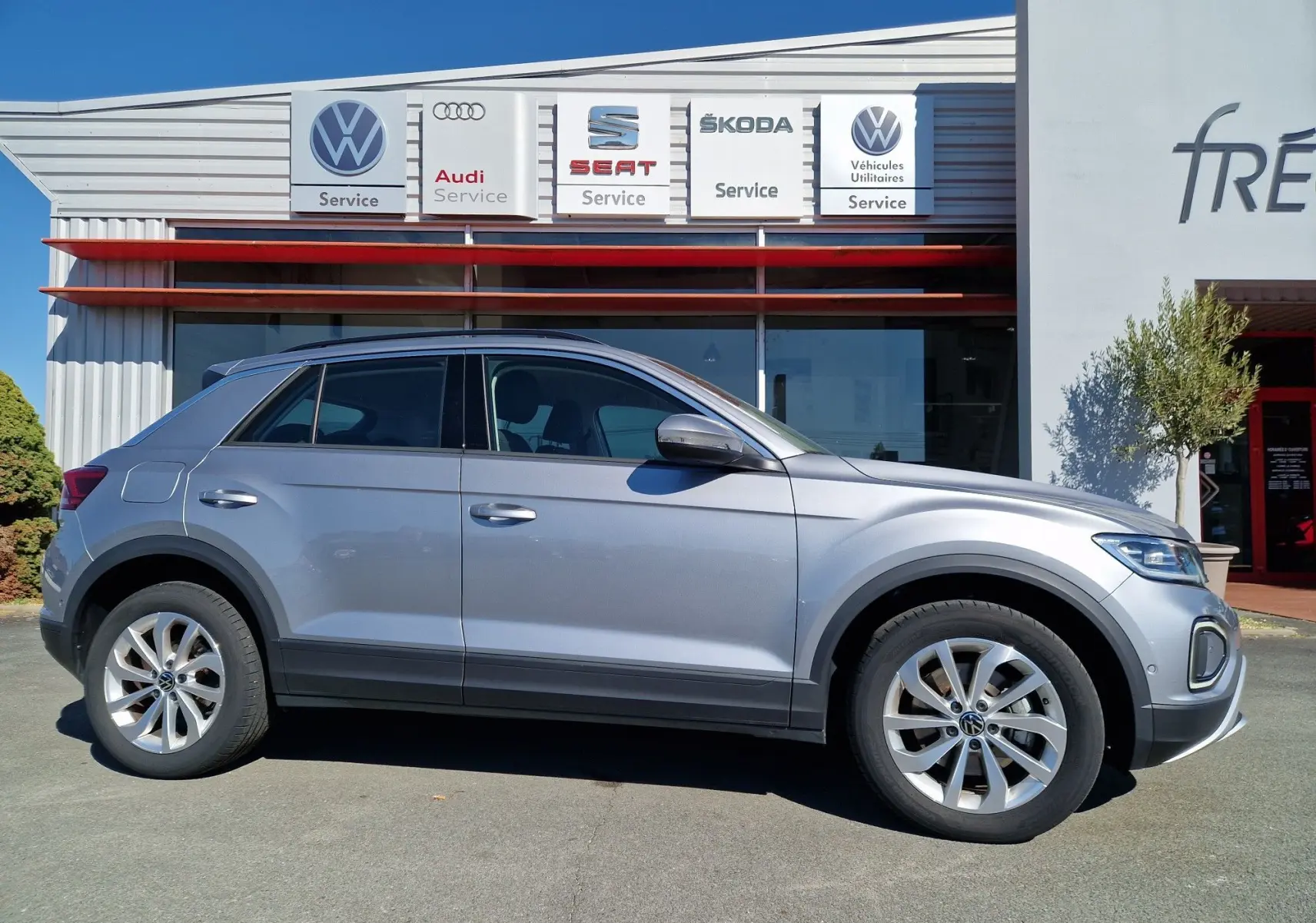 Profil côté gauche d'un Volkswagen T-Roc gris pyrite 2023 garé devant un centre de service multimarques.