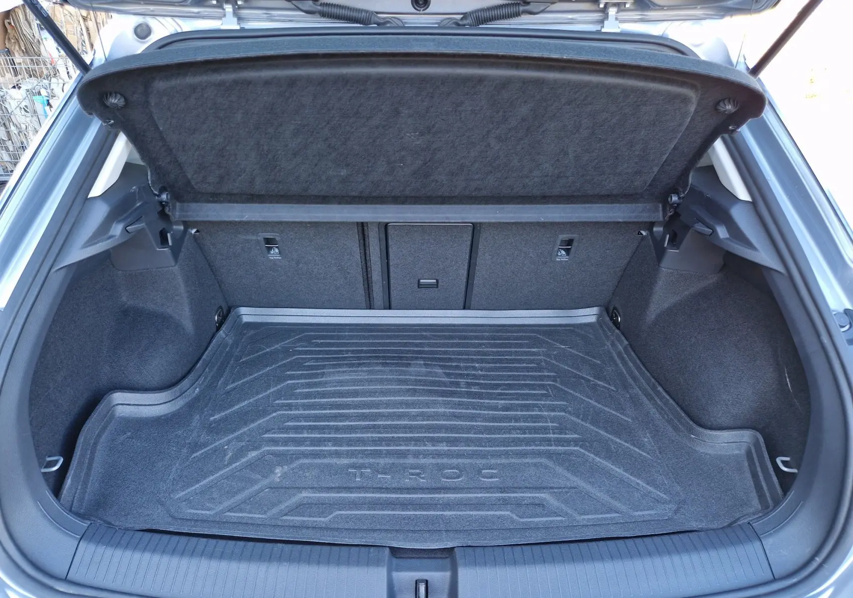 Coffre ouvert vu de l'arrière d'un Volkswagen T-Roc gris Pyrite, avec tapis de coffre marqué T-ROC.