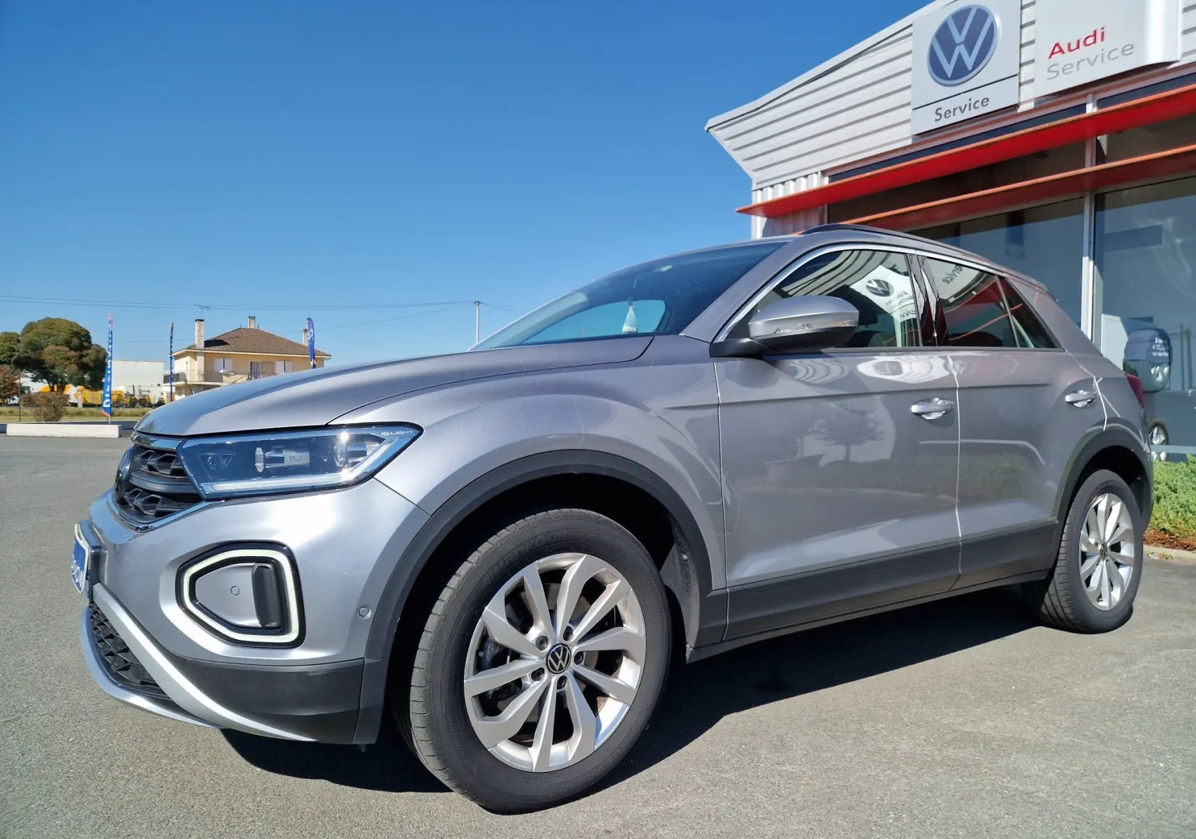 Volkswagen T-Roc gris pyrite en 3/4 avant droit, stationné devant un service Volkswagen et Audi par temps clair.