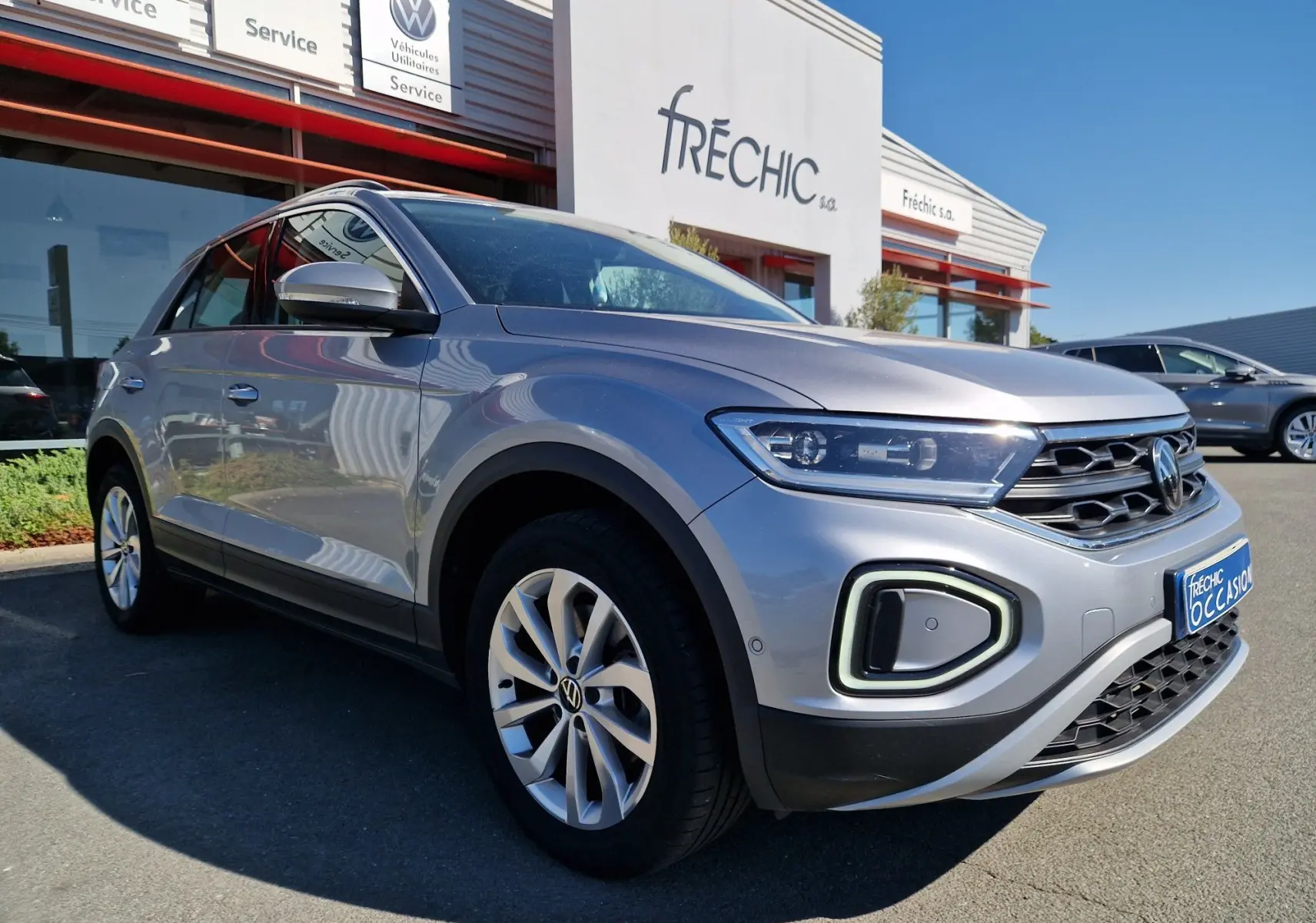 Volkswagen T-Roc gris pyrite vue 3/4 avant droit, avec phares LED et calandre distinctive, stationné devant un concessionnaire.