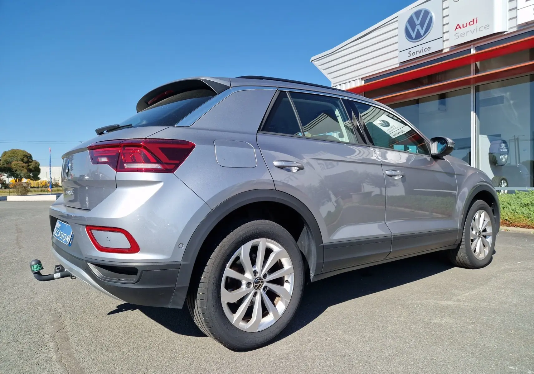 Volkswagen T-Roc gris pyrite vu 3/4 arrière droit avec attelage, stationné devant un service Volkswagen.
