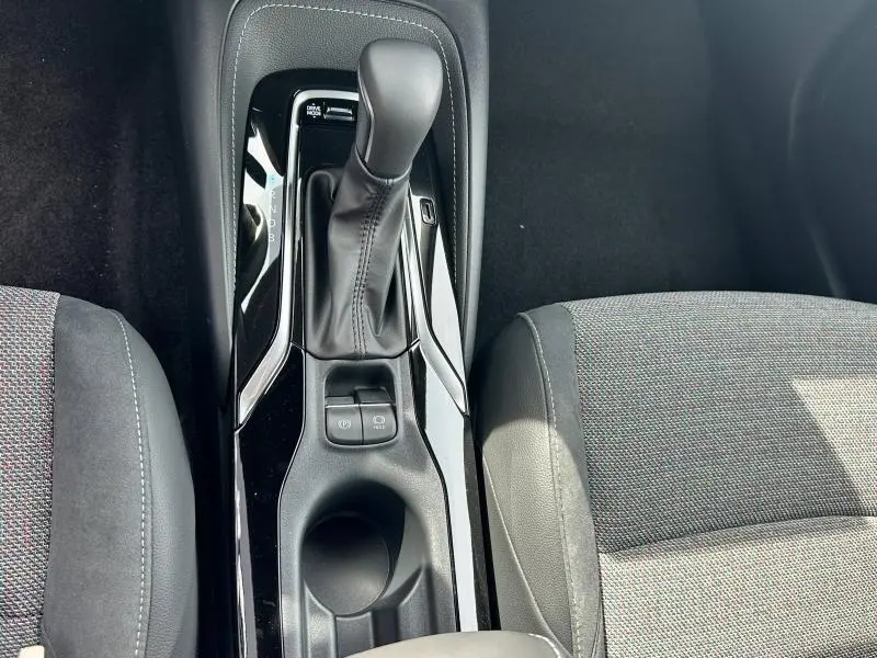 Vue plongeante sur la console centrale noire de la Toyota Corolla 1.8 Hybrid 2025 avec levier de vitesse automatique et sièges tissu noir.