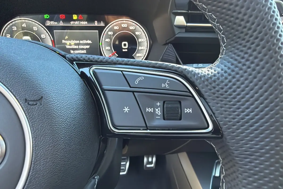 Gros plan sur les commandes au volant à méplat de l'Audi A3 TFSI 150 Hybrid S-Line 2025 avec tableau de bord numérique.